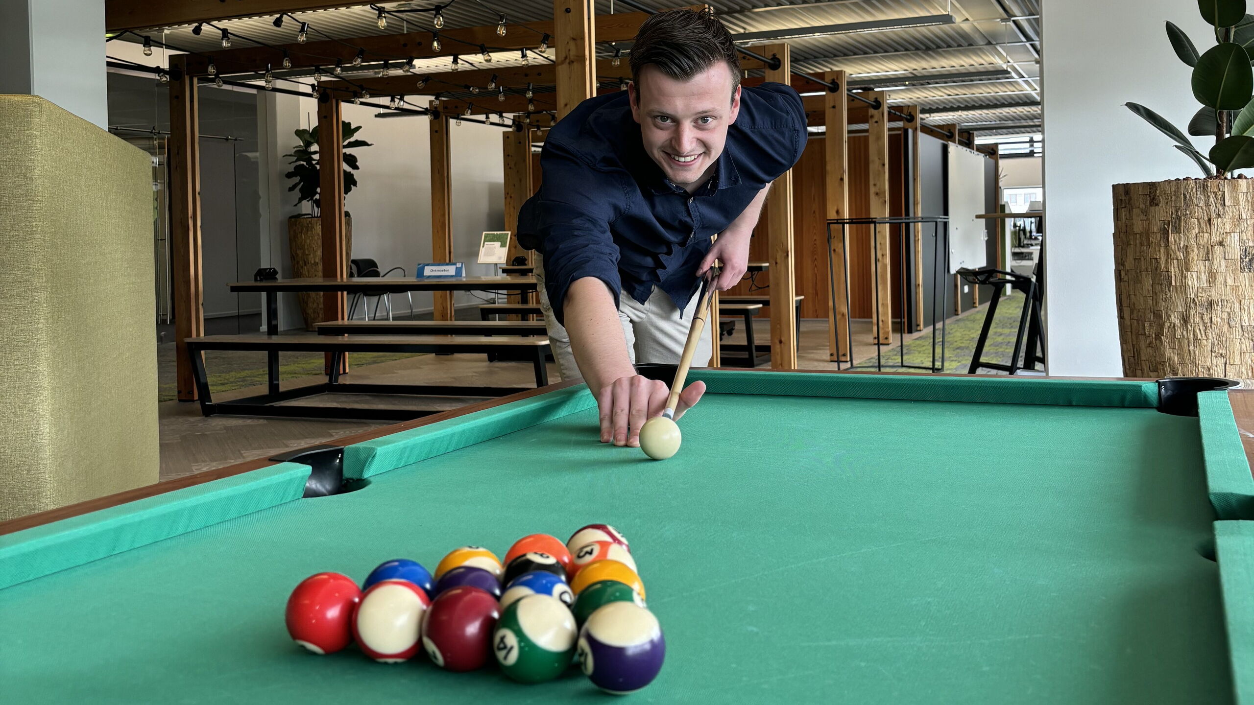 Collega Tim stoot de witte bal aan op een biljarttafel in een kantooromgeving, klaar om het spel te openen.+