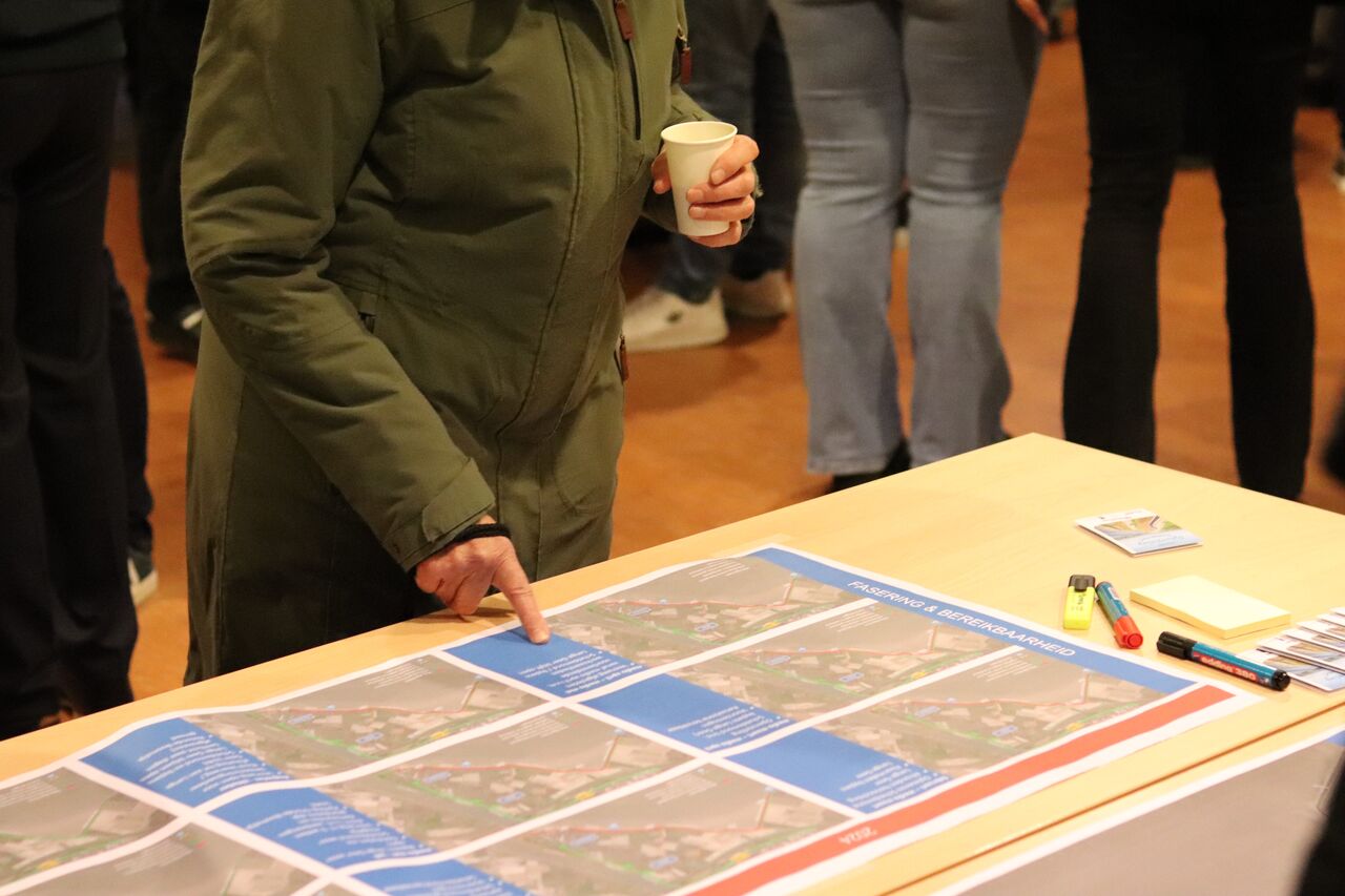 weergave van de inloopavond. bezoeker in het dorpshuis kijkt en wijst naar de tekeningen. 