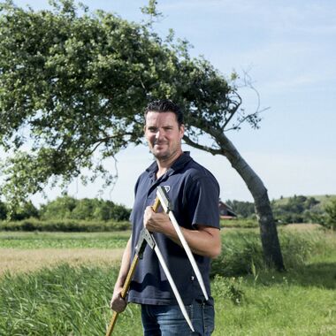 Man staat in een weiland voor een boom met een meetapparaat in zijn handen.